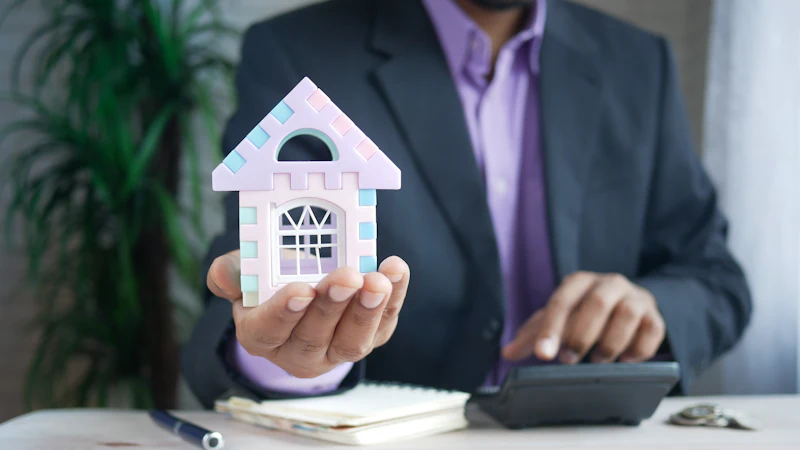 A person at a desk holding a model house next to a calculator and paperwork