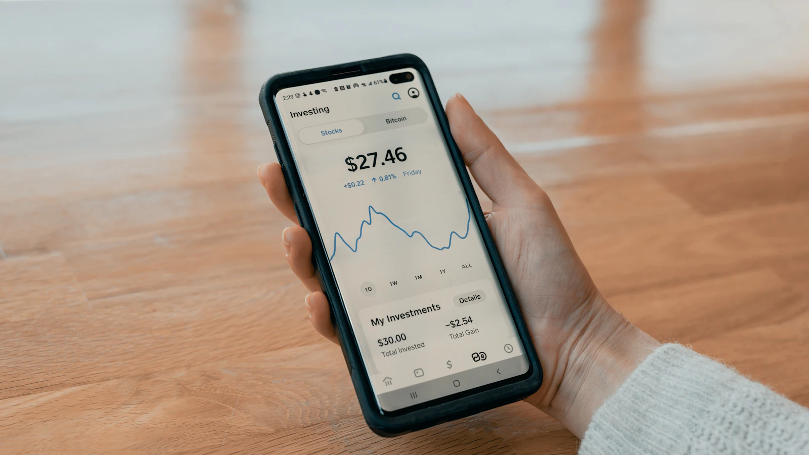A hand holding a smartphone at a wooden table, showing a personal investing app with a balance and rising line chart
