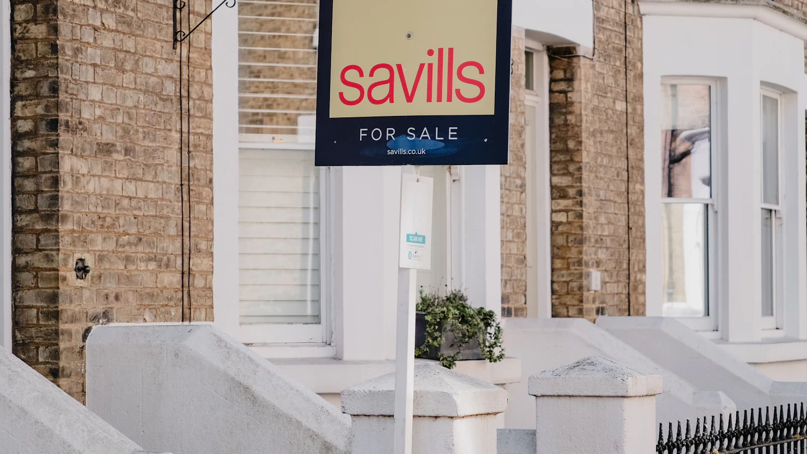 A Savills "For Sale" sign mounted outside a row of brick Victorian townhouses in the UK